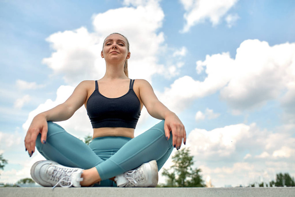 Beautiful athletic girl in lotus pose against the sky in sportswear, element of yoga. Sport motivation, copy space, banner for advertising, think wider. - Foto, Imagen