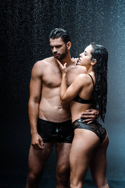 sexy and wet girl touching face of shirtless man standing under raindrops on black  - Fotografie, Obrázek