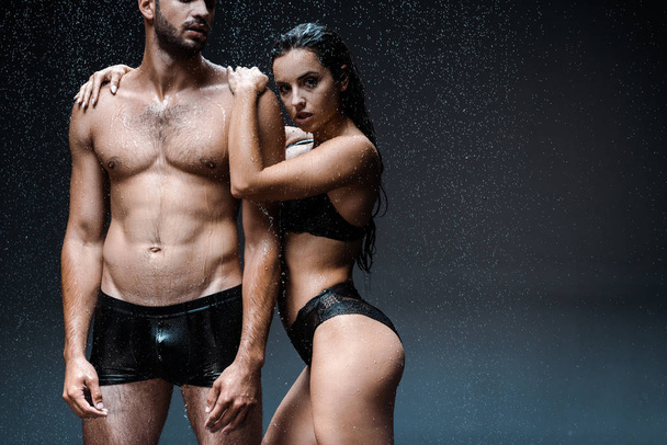 vista incrociata di uomo senza maglietta in piedi con ragazza sexy e bagnata sotto gocce di pioggia sul nero
  - Foto, immagini