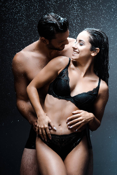 feliz sin camisa hombre abrazando alegre mojado chica bajo gotas de lluvia en negro
  - Foto, Imagen