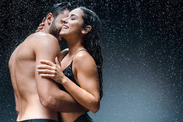 黒の雨滴の下で幸せなガールフレンドを抱きしめる濡れシャツレスの男  - 写真・画像