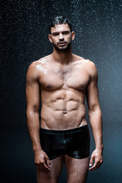 wet handsome man looking at camera while standing under raindrops on black  - Foto, immagini