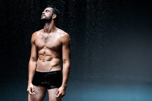 hombre sin camisa y mojado con los ojos cerrados de pie bajo gotas de lluvia en negro
  - Foto, Imagen