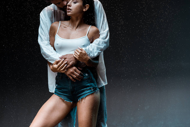 przycięte widok człowieka przytulanie sexy mokrej dziewczyny pod krople deszczu na czarnym  - Zdjęcie, obraz