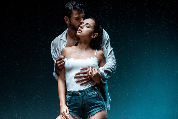 handsome man standing and kissing sexy girlfriend with closed eyes under raindrops on black  - 写真・画像