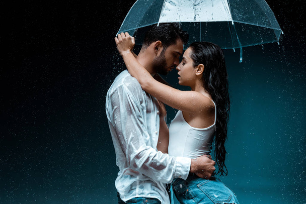 Seitenansicht der attraktiven Freundin umarmt gutaussehenden Freund und hält Regenschirm auf schwarz  - Foto, Bild