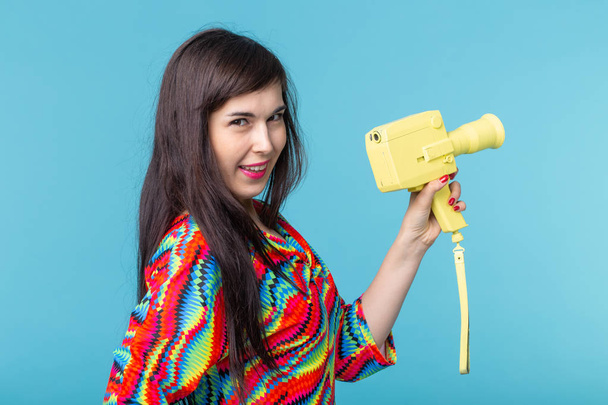 Portrait of a smiling young woman with a vintage yellow video camera in her hands posing against a blue background. Concept of video and photography - Foto, Imagem
