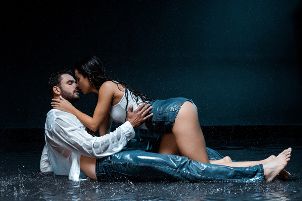 sexy and wet woman touching face of handsome boyfriend lying under raindrops on black  - Photo, Image