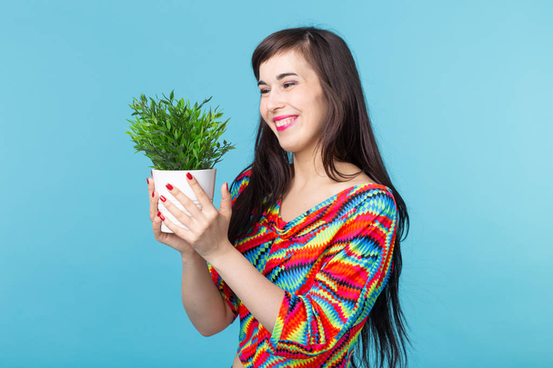 Iloinen nuori nainen pitelee ruukkua, jossa kasvi poseeraa sinistä taustaa vasten. Käsitys puutarhanhoito ja sisustustarvikkeet
. - Valokuva, kuva
