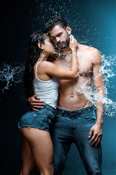 sexy a vlhká žena, která objímá hezkého a svalnatého přítele pod dešťovou kapek na černém  - Fotografie, Obrázek