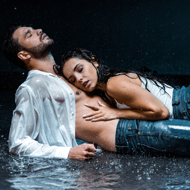 sexy en natte meid liggend met knappe vriendje onder regendruppels op zwart  - Foto, afbeelding