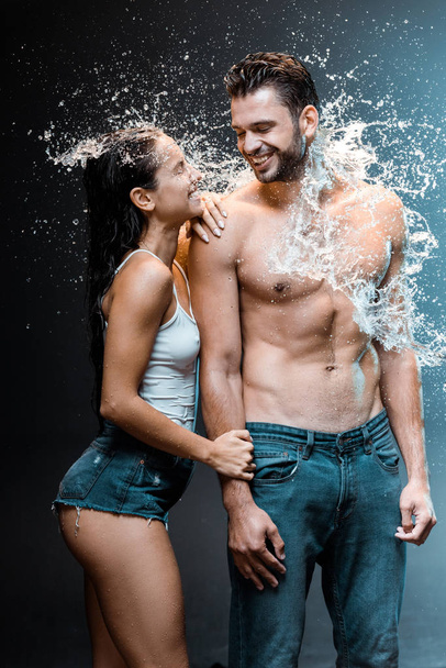shirtless man glimlachend met aantrekkelijke vriendin in denim shorts in de buurt van splash van water op zwart  - Foto, afbeelding