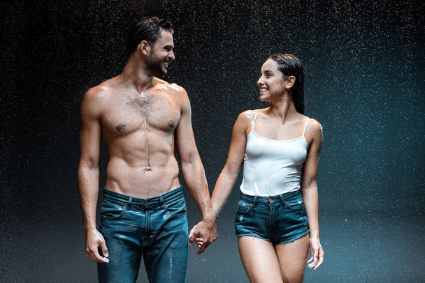 homem sem camisa feliz de mãos dadas com a menina molhada em shorts jeans em preto
  - Foto, Imagem