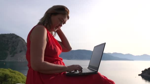 Tired woman working at a laptop and yawning sitting against a sea landscape - Footage, Video