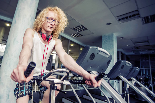 Gym. Skinny blond guy with glasses and red headphones trains on  - Фото, изображение