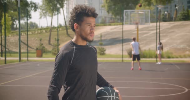 Closeup portrait of young handsome african american male basketball player holding ball and looking at camera outdoors - Filmagem, Vídeo