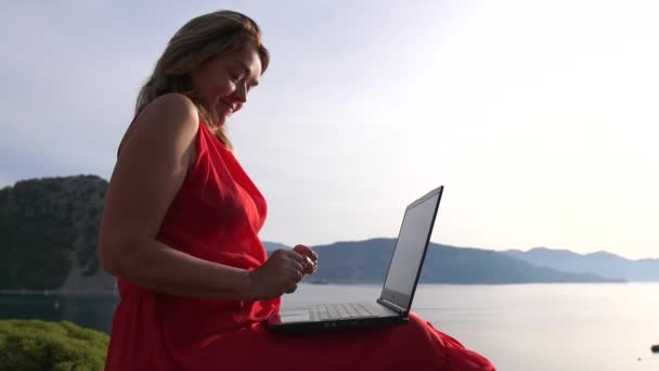 Mulher sorrindo digitando no laptop sentado contra uma bela paisagem marinha, câmera lenta
 - Filmagem, Vídeo