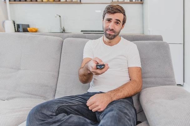Beautiful hilarious young man holding a remote control. During this, the TV is watching while sitting on the couch at home. - Fotografie, Obrázek