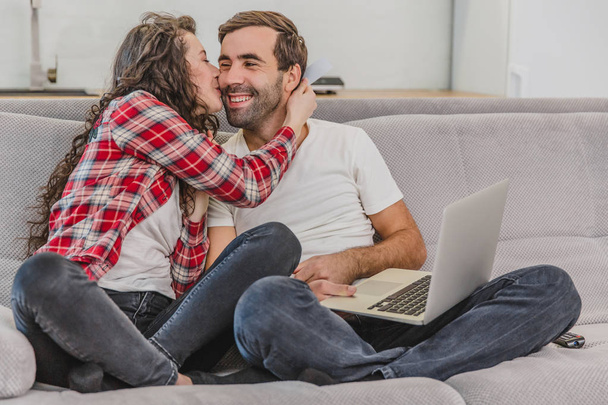 Smiling young couple, enjoying the time spent at the computer, sitting on the couch at home together, happy man, the woman kisses her husband on the cheek. - Foto, Bild