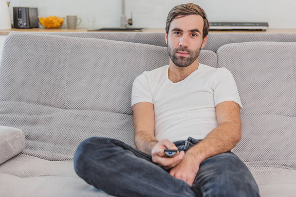リモートコントロールを持っている美しい陽気な若い男。この間に、テレビは家でソファーに座っている間に見ている. - 写真・画像