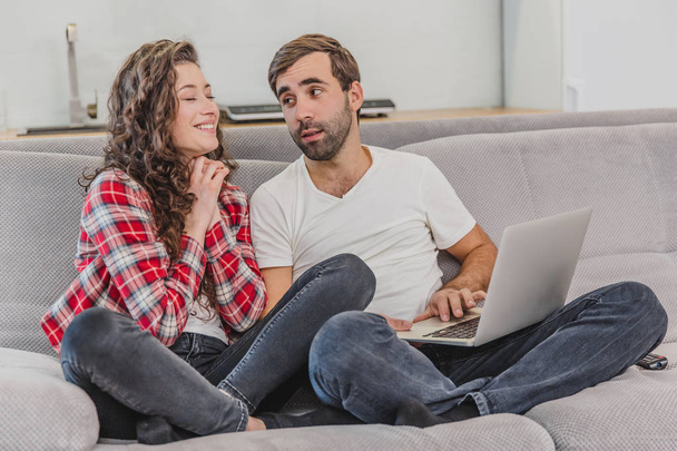 A good couple while shopping online using a laptop and smiling while sitting at a couch at home. A woman asks her husband to make her purchase online. - 写真・画像