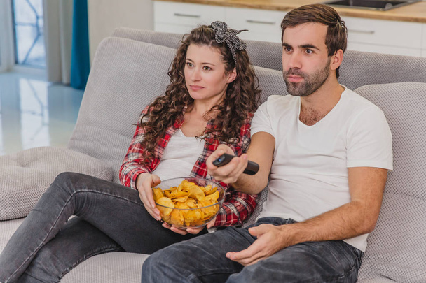 Smiling wife and man sitting on the couch. Watch the TV while eating chips in bed. - 写真・画像