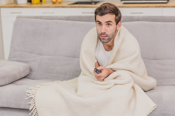 A man with a cold sitting on the couch in a blanket is as cold as possible. Man at home - sick man. Gray sofa in the room. The concept of a misfortune. The concept of health and healthy lifestyle. - Photo, Image