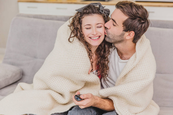 junges Paar, das es sich in seiner Wohnung gemütlich macht, auf der Couch sitzt und fernsieht. bedeckt mit einer Decke, glückliche Umarmung. - Foto, Bild