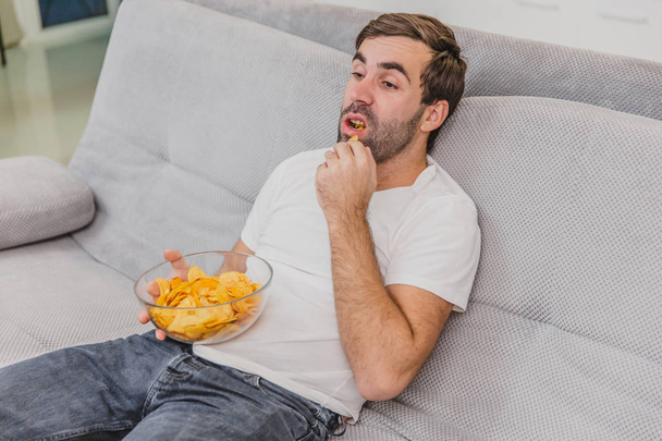 The guy in the T-shirt sits on the couch, eats chips and watches the TV. While holding the remote in his hands. Personality at home. - Zdjęcie, obraz
