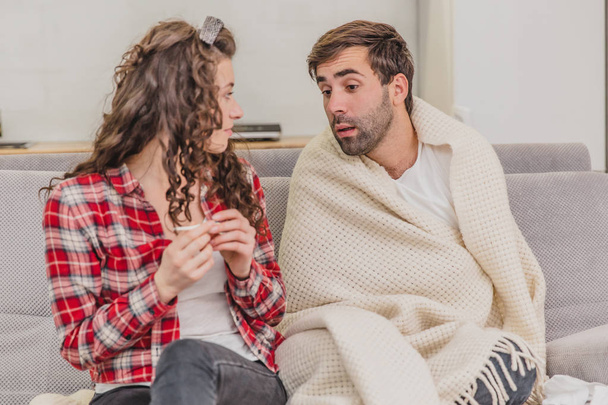A man with a cold sitting on a sofa in a blanket blanket. Checking the temperature. The wife is sitting next to her and helps. Man at home. Sick young man Gray sofa in the room. Unhappy guy. Concept - Foto, Imagen