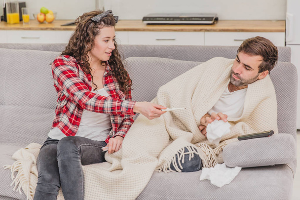 A man with a cold sitting on a sofa in a blanket blanket. Checking the temperature. The wife is sitting next to her and helps. Man at home. Sick young man Gray sofa in the room. Unhappy guy. Concept - Foto, immagini