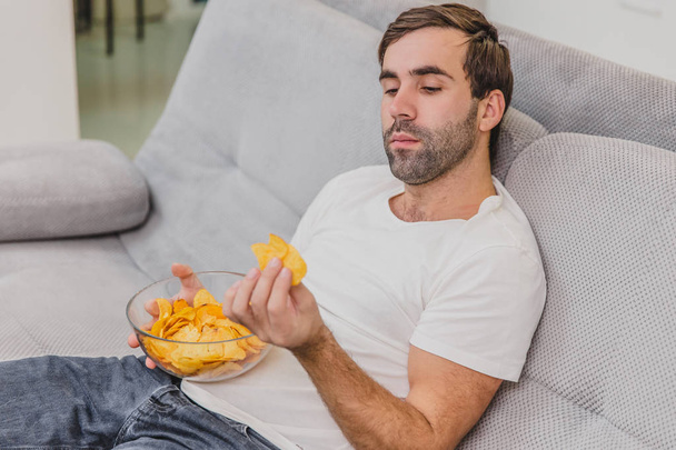 The guy in the T-shirt sits on the couch, eats chips and watches the TV. While holding the remote in his hands. Personality at home. - Foto, Imagem
