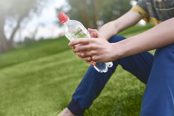 junger kaukasischer Läufer entspannt mit Trinkwasserflasche in der Hand und sitzt nach Sport am frühen Morgen auf dem Rasen im Park, Bewegung und gesundes Konzept. - Foto, Bild