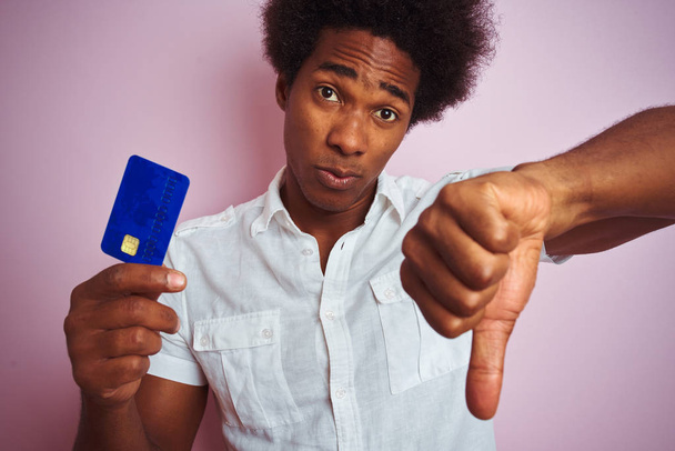 Afro-amerikai vevő ember Holding hitelkártya állt át elszigetelt rózsaszín háttér dühös arc, negatív jel mutatja nem tetszik a hüvelykujját lefelé, elutasítás koncepció - Fotó, kép
