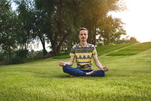 Νέος άνθρωπος που διαλογίζεται σε εξωτερικούς χώρους στο πάρκο, καθισμένος με τα μάτια κλειστά και τα χέρια του ενωμένα. απολαμβάνοντας τη φύση, τη γιόγκα και την αντίληψη του διαλογισμού. πνευματικές πρακτικές. Zen - Φωτογραφία, εικόνα