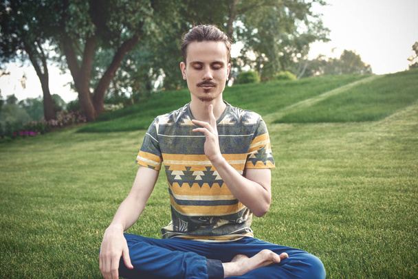 Young man meditating outdoors in the park, sitting with eyes closed and his hands together.enjoying nature, yoga and meditation concept. spiritual practices.zen - Photo, Image