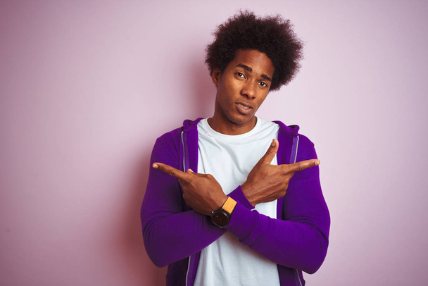 孤立したピンクの背景の上に立つ紫色のスウェットシャツを着た若いアフリカ系アメリカ人男性指で両側を指差し、異なる方向は反対 - 写真・画像