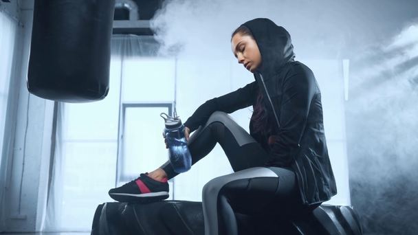 beautiful sportswoman in hoodie sitting on tire, looking at camera and drinking water in smoky gym - Video
