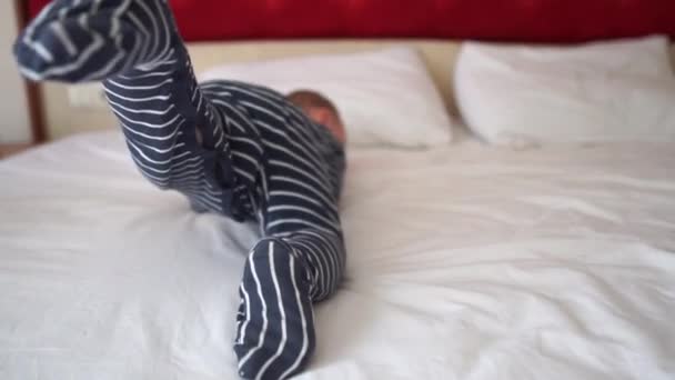 ストライプのジャンプスーツを着た小さな男の子がホテルの大きなベッドで回転している、スローモーション - 映像、動画