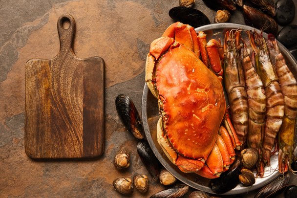 top view of uncooked crab, shellfish, cockles and mussels in bowl near wooden chopping board on textured surface  - Fotografie, Obrázek