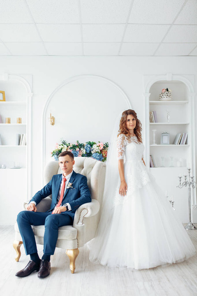 Beautiful bride in white dress and groom in suit, posing in white Studio interior, wedding - Фото, изображение