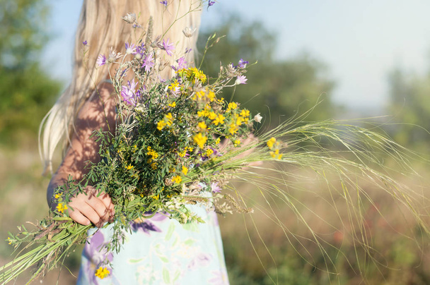 Kis aranyos szőke lány egy területen egy napsütéses napon egy csokor vadvirág.  - Fotó, kép
