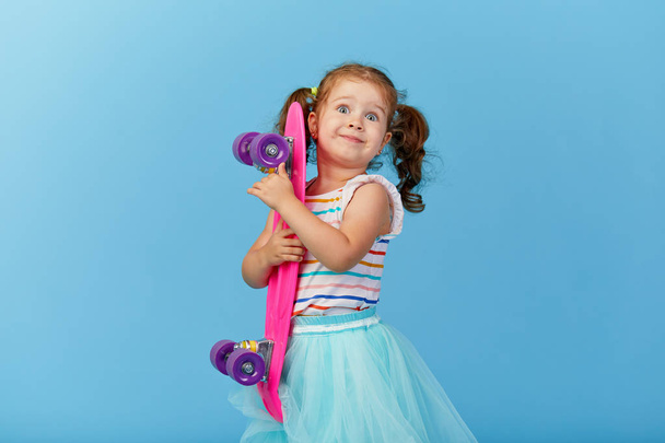 Cool little toddler girl in bright clothes holds a skateboard and looking at the camera isolated on blue background - Foto, Bild