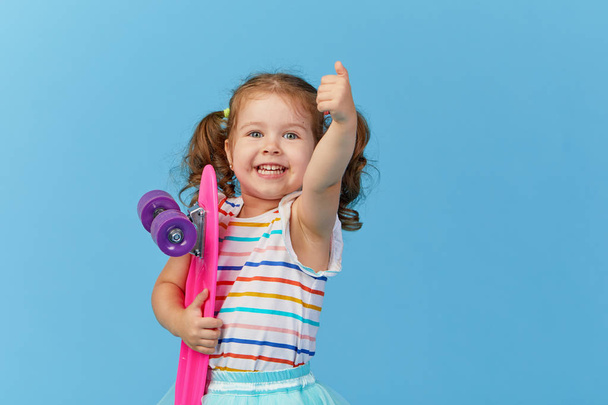 Cool little toddler girl in bright clothes holds a skateboard and looking at the camera isolated on blue background. Shows thumb up - Fotó, kép