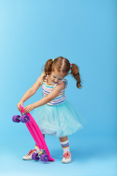 Cool little toddler girl in bright clothes holds a skateboard and looking at the camera isolated on blue background - 写真・画像