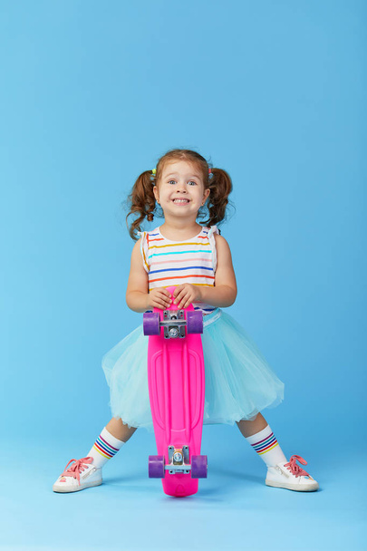 Cool little toddler girl in bright clothes holds a skateboard and looking at the camera isolated on blue background - Foto, immagini