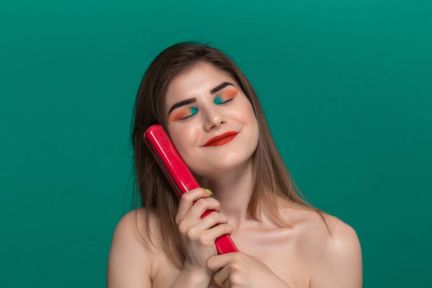 Portré gyönyörű fiatal nő, élénk színű make-up csinál frizurát magát egy lila egyengető vas. - Fotó, kép