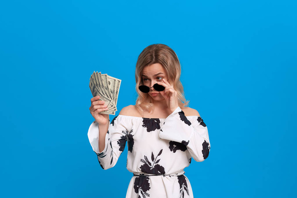 Portrait of a girl with curly blond hair in a white dress and dark sunglasses standing on a blue background looking surprisingly at the bundle of dollars. - Photo, Image