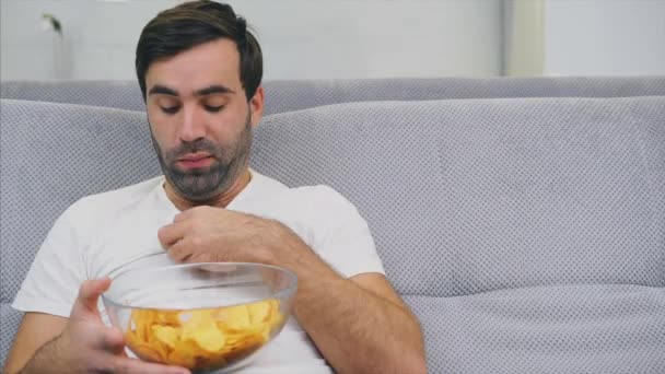 The guy in the T-shirt sits on the couch, eats chips and watches the TV. While holding the remote in his hands. - Felvétel, videó