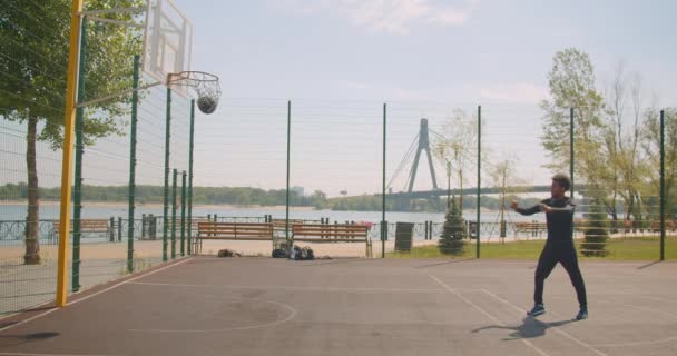 Portrait of sporty cheerful african american male basketball player throwing a ball into a hoop and celebrating with triumph on the court in the urban city outdoors with bridge on the background - Materiaali, video